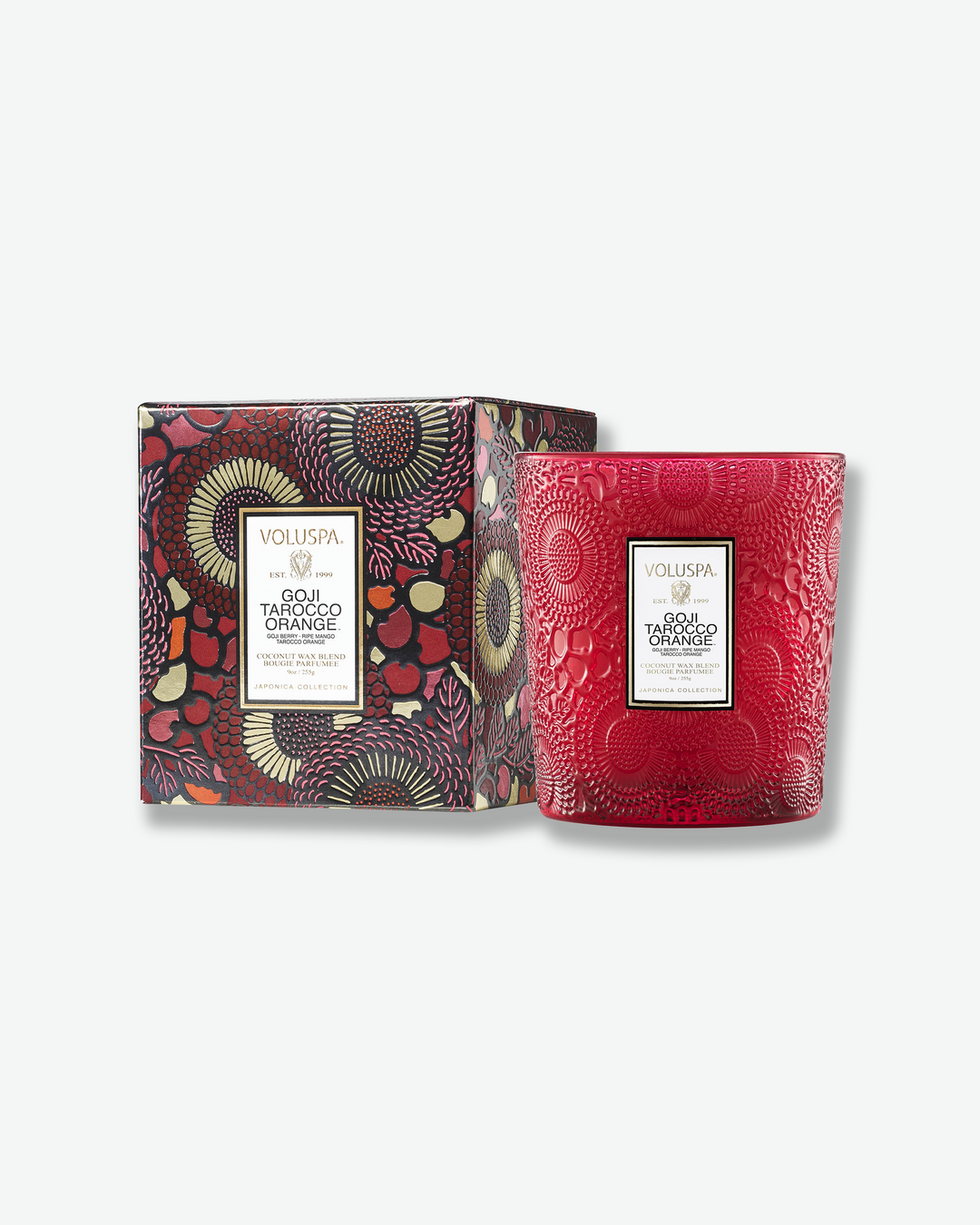 High-angle product shot of a burning Voluspa Goji Tarocco Orange textured glass candle against a plain gray background. The single-wick flame glows brightly from the center of the pool of melted white wax inside the translucent, embossed red glass holder.