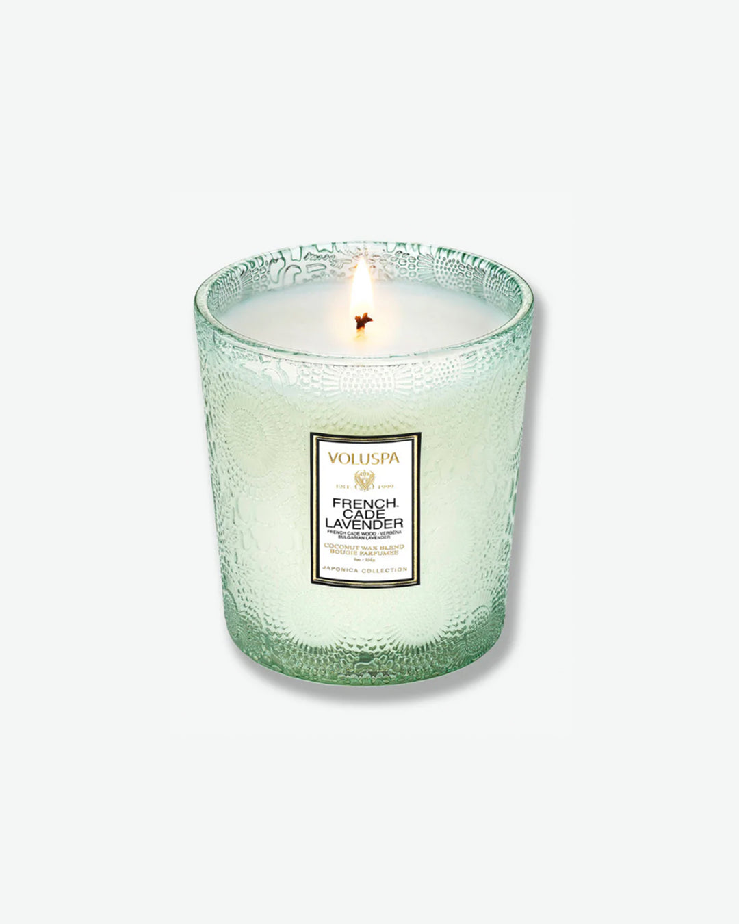 High-angle product shot of a burning Voluspa French Cade Lavender textured glass candle against a plain gray background. The single-wick flame burns in the center of the pale green, embossed glass holder, illuminating the melted wax.