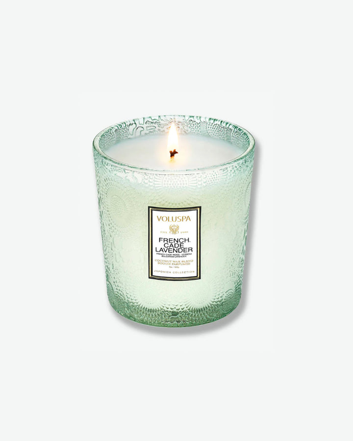 High-angle product shot of a burning Voluspa French Cade Lavender textured glass candle against a plain gray background. The single-wick flame burns in the center of the pale green, embossed glass holder, illuminating the melted wax.