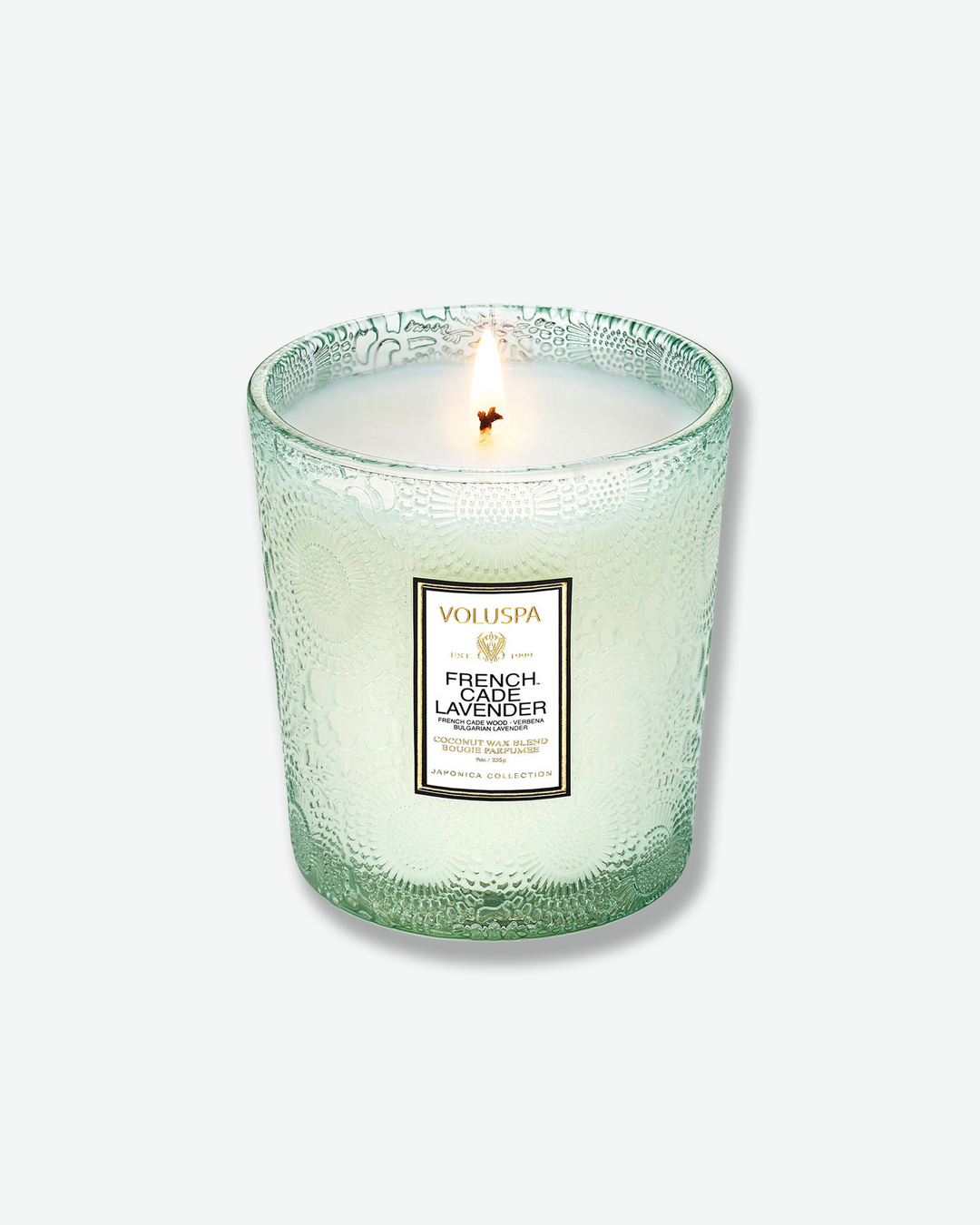 High-angle product shot of a burning Voluspa French Cade Lavender textured glass candle against a plain gray background. The single-wick flame burns in the center of the pale green, embossed glass holder, illuminating the melted wax.