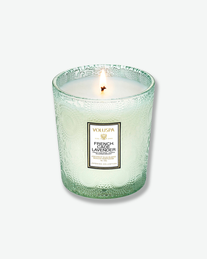 High-angle product shot of a burning Voluspa French Cade Lavender textured glass candle against a plain gray background. The single-wick flame burns in the center of the pale green, embossed glass holder, illuminating the melted wax.