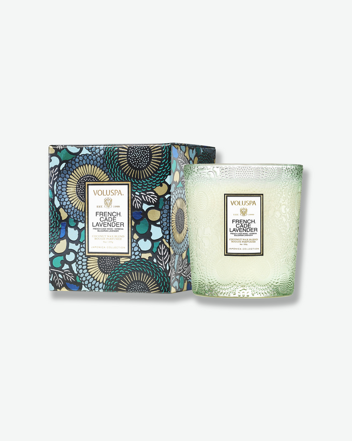 Product shot of a Voluspa French Cade Lavender classic textured glass candle standing next to its matching square outer packaging. The light green glass candle jar is detailed with an intricate embossed floral motif and sits beside a box featuring a blue, green, and black botanical pattern.