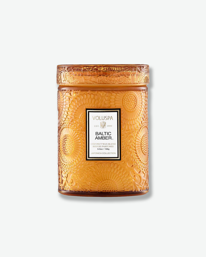 Front view of a Voluspa Baltic Amber small jar candle against a plain gray background. The warm, amber-colored glass vessel is completely detailed with an embossed floral pattern and capped with a matching textured glass screw-on lid.