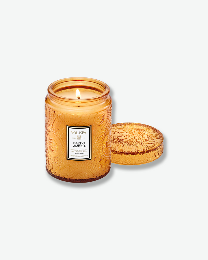 Product shot of a lit Voluspa Baltic Amber small glass jar candle. The single wick burns inside the amber-colored glass vessel, which features an intricate embossed floral motif. The matching textured amber glass lid has been removed and sits flat beside the candle.