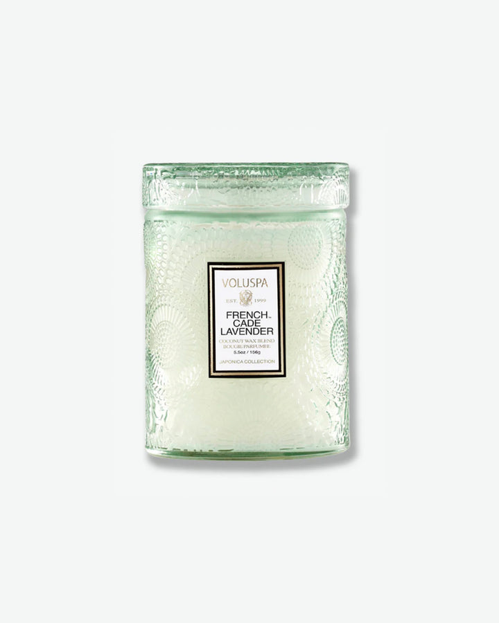 Front view of a Voluspa French Cade Lavender small jar candle against a plain gray background. The candle is housed in a translucent, pale sage green glass container embossed with a detailed botanical pattern and features a central white label, topped with a matching textured glass lid.