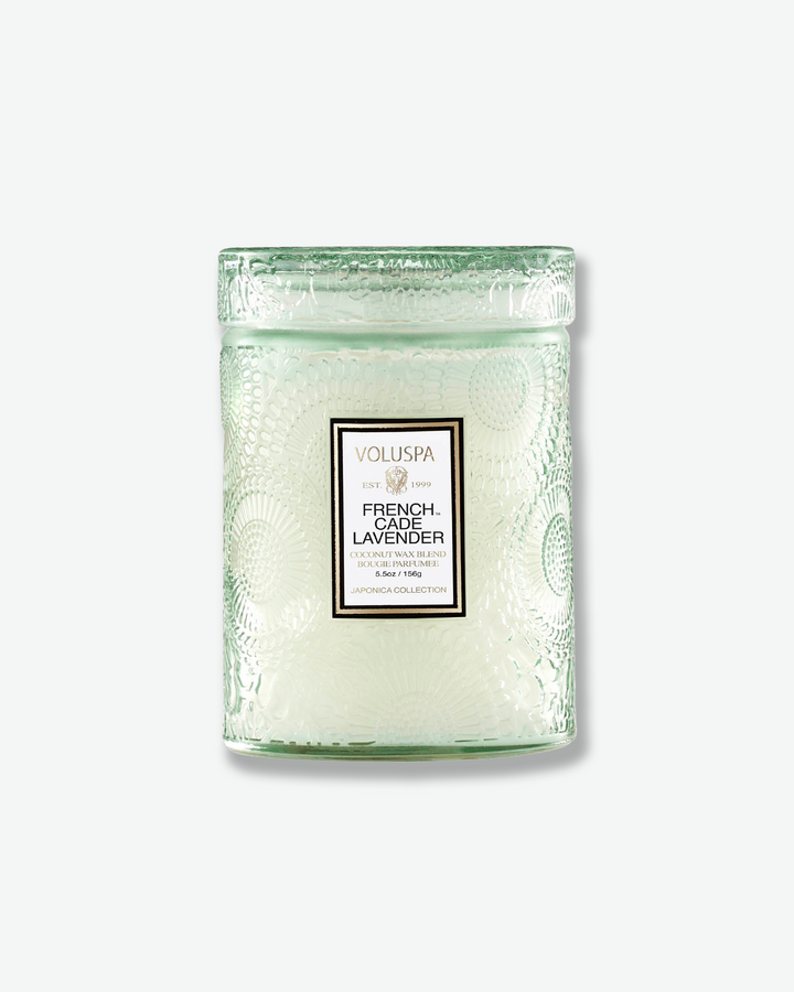 Front view of a Voluspa French Cade Lavender small jar candle against a plain gray background. The candle is housed in a translucent, pale sage green glass container embossed with a detailed botanical pattern and features a central white label, topped with a matching textured glass lid.