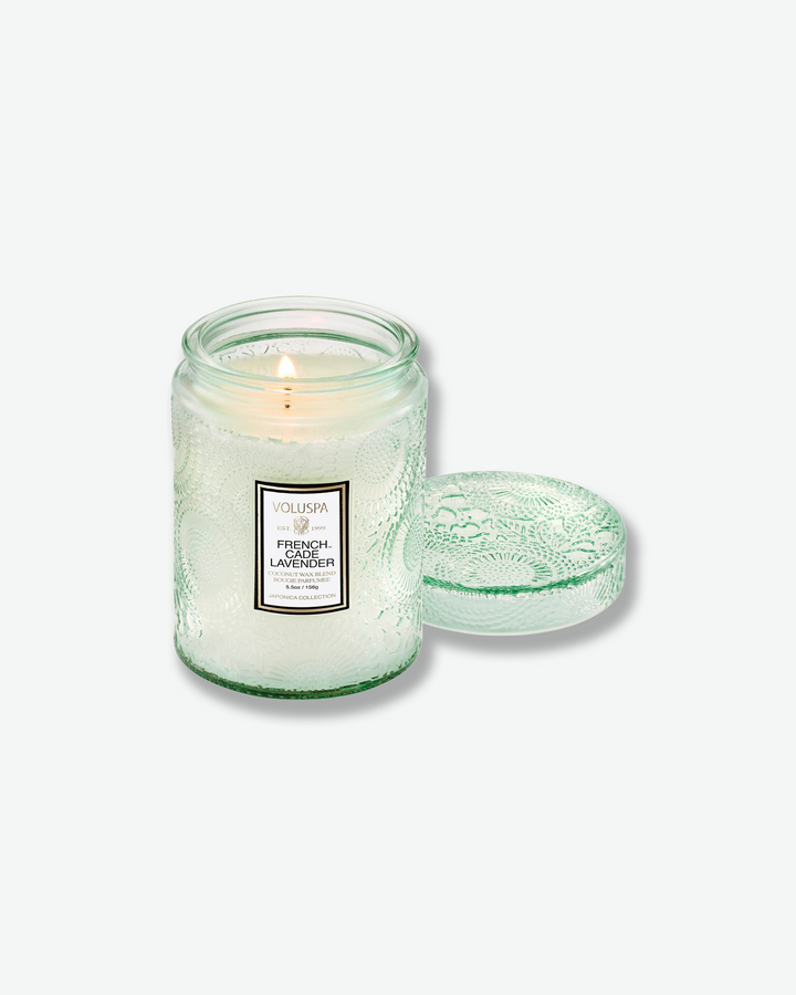 Product shot of a lit Voluspa French Cade Lavender small glass jar candle. A single-wick flame glows within the pale green embossed jar, and its matching textured glass lid rests flat on the surface to the right.