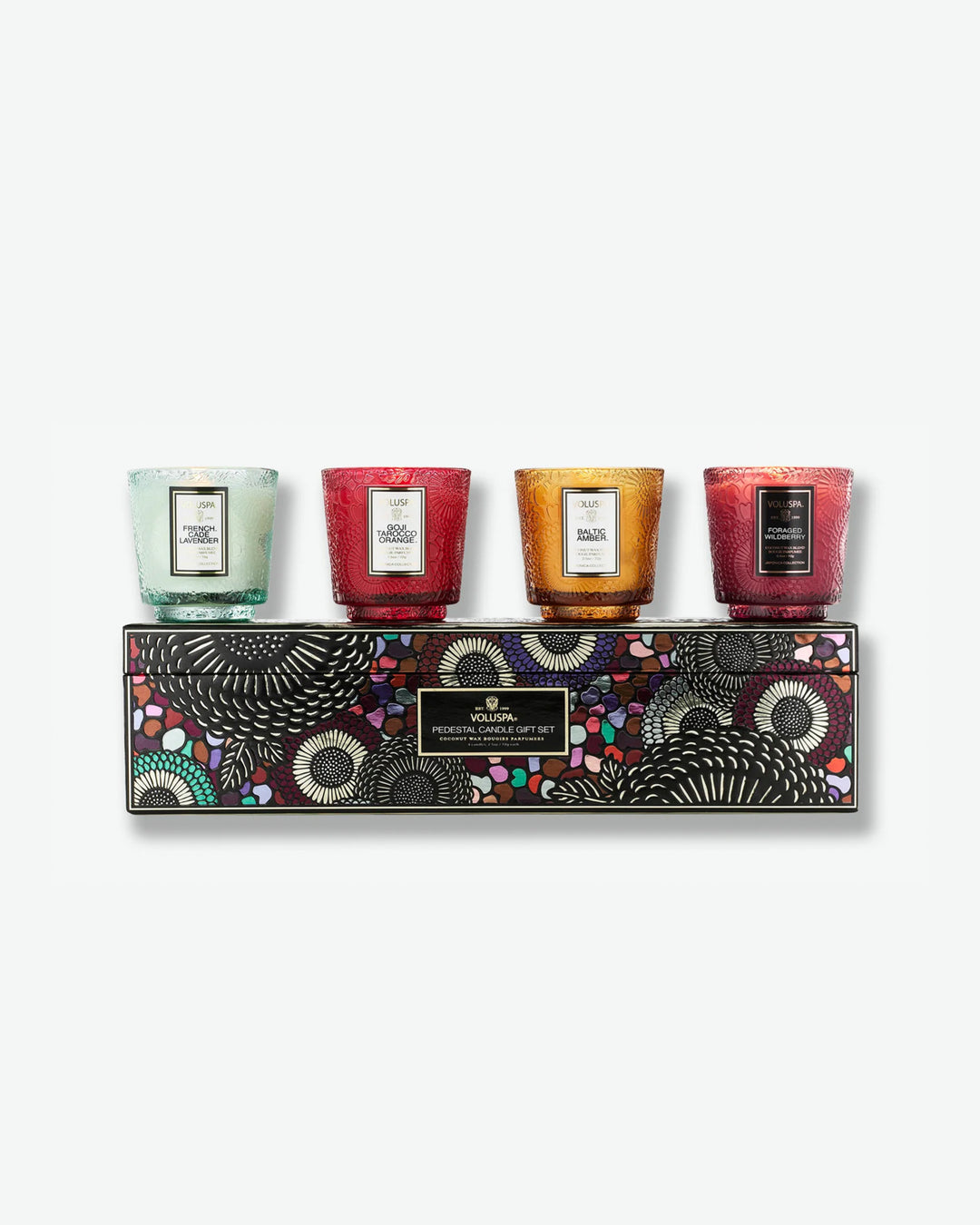 Product shot showcasing the Voluspa Pedestal Candle Gift Set. Four small, colorful, pedestal-style textured glass candles—mint green, ruby red, amber gold, and berry purple—are lined up on top of a rectangular display box featuring a vibrant, multi-colored Japonica floral pattern.
