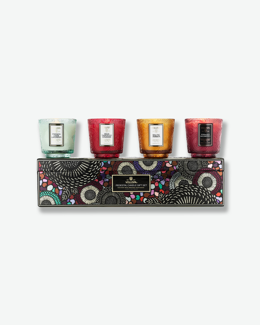 Product shot showcasing the Voluspa Pedestal Candle Gift Set. Four small, colorful, pedestal-style textured glass candles—mint green, ruby red, amber gold, and berry purple—are lined up on top of a rectangular display box featuring a vibrant, multi-colored Japonica floral pattern.