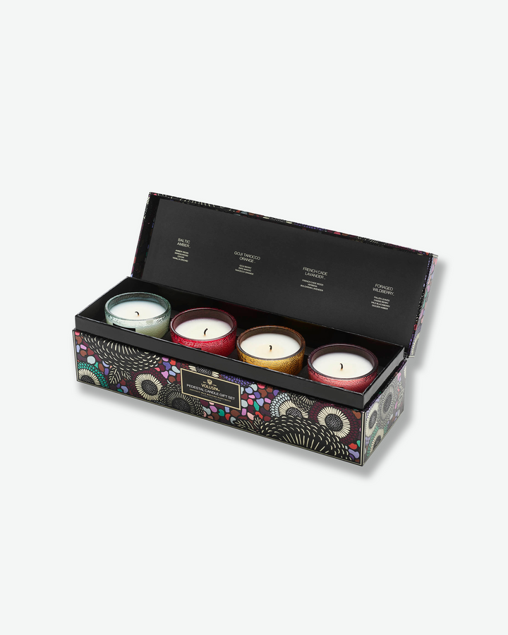 Three-quarter angled view of the open Voluspa Pedestal Candle Gift Set box. The patterned lid is flipped up to reveal four distinct, unlit pedestal glass candles nestled securely in a row within the black velvet-lined interior tray.
