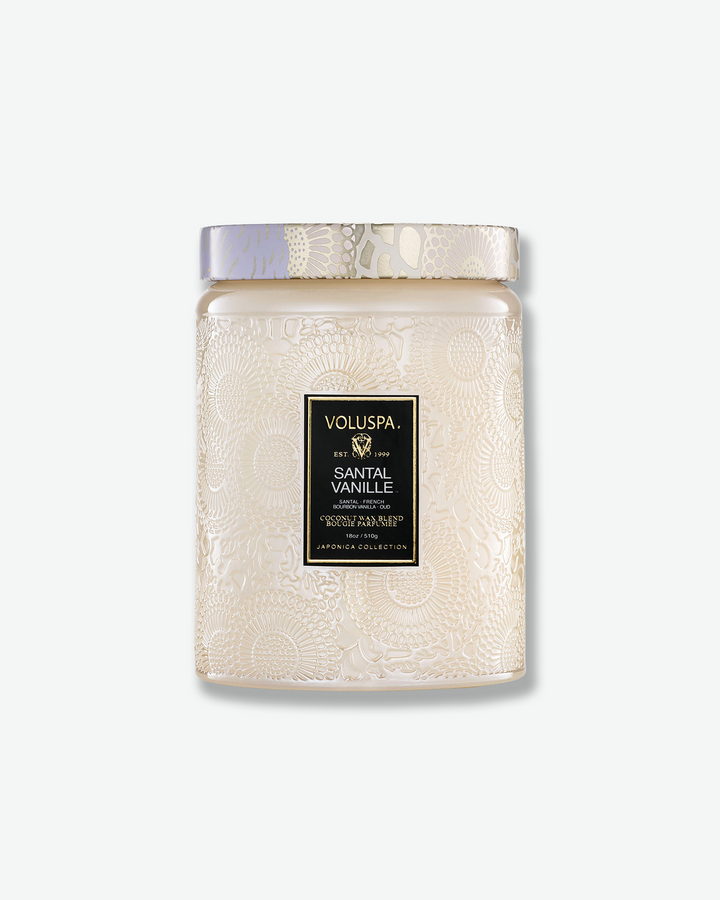 Front view of a Voluspa Santal Vanille large jar candle against a plain gray background. The candle is housed in an elegant, cream-colored translucent glass jar featuring an intricate embossed floral motif, a central product label with a gold border, and a matching patterned tin lid.