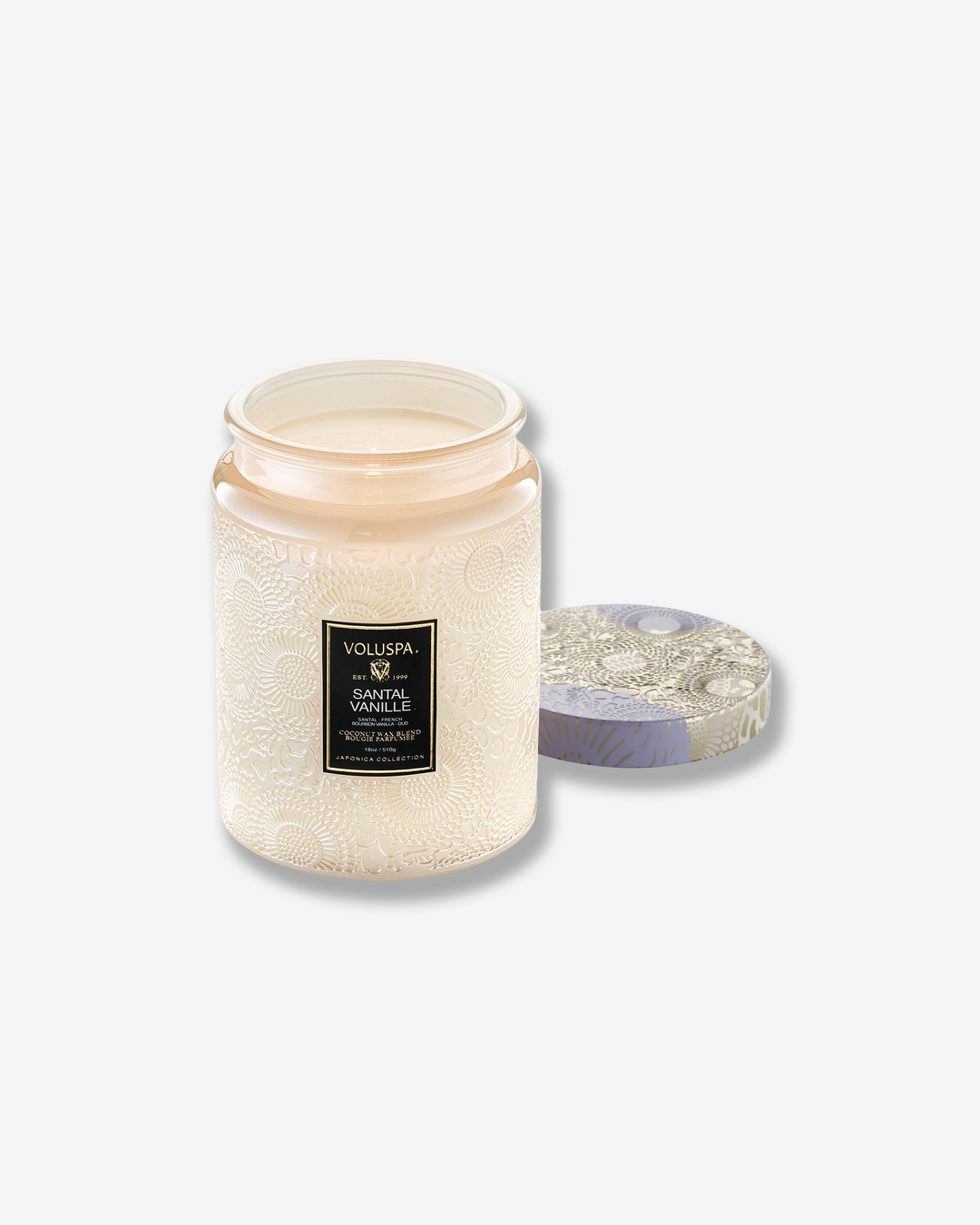 Product shot of a lit Voluspa Santal Vanille large jar candle. A single flame burns smoothly inside the cream-tinted embossed glass container, with the decorative muted purple and cream tin lid resting face-up on the surface next to the jar.