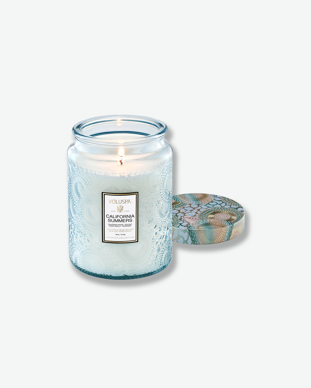 Product shot of a lit Voluspa California Summers large jar candle. The single wick glows warmly within the light blue embossed glass jar, and the removed decorative tin lid sits flat to the right of the candle.
