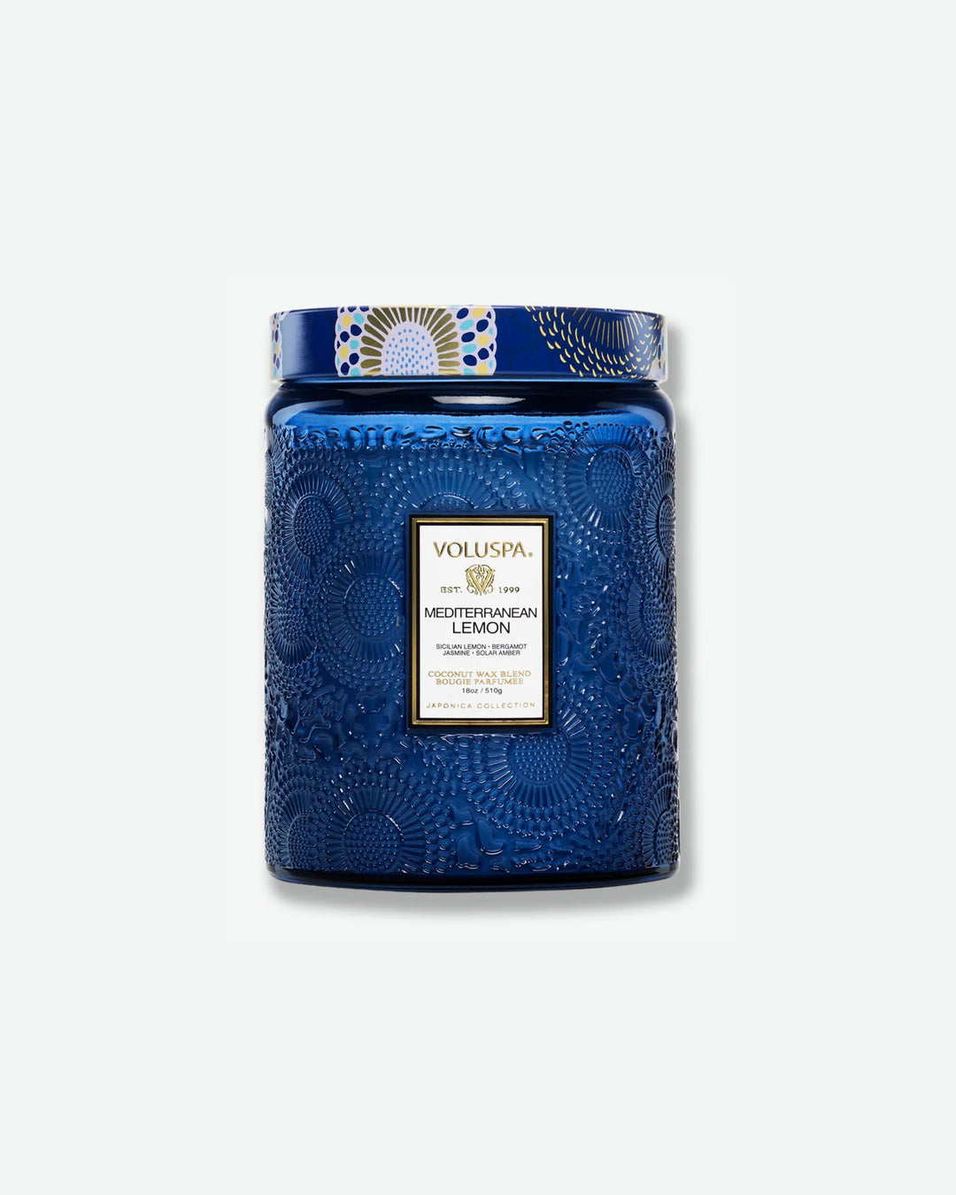 Front view of a Voluspa Mediterranean Lemon large jar candle against a plain gray background. The candle features a deep cobalt blue glass jar intricately embossed with swirling floral textures, finished with a black and gold central label and a decorative blue, yellow, and white patterned tin lid.