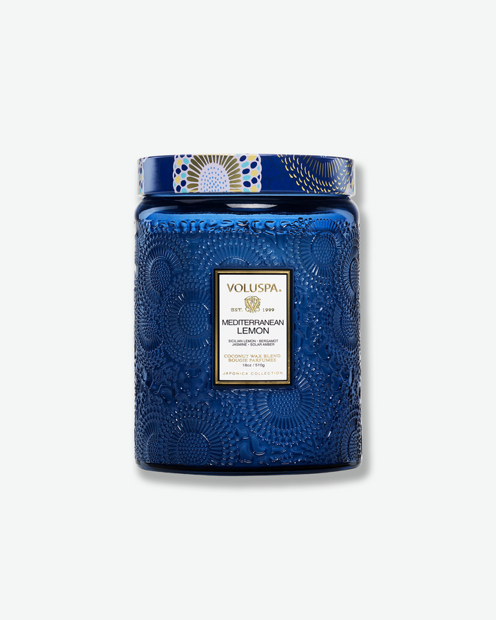 Front view of a Voluspa Mediterranean Lemon large jar candle against a plain gray background. The candle features a deep cobalt blue glass jar intricately embossed with swirling floral textures, finished with a black and gold central label and a decorative blue, yellow, and white patterned tin lid.