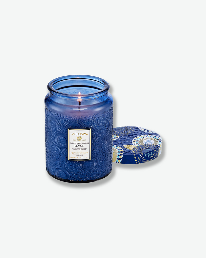 Product shot of a lit Voluspa Mediterranean Lemon large jar candle. The flame burns gently inside the cobalt blue embossed glass vessel, with its matching patterned blue tin lid resting flat on the surface next to it.