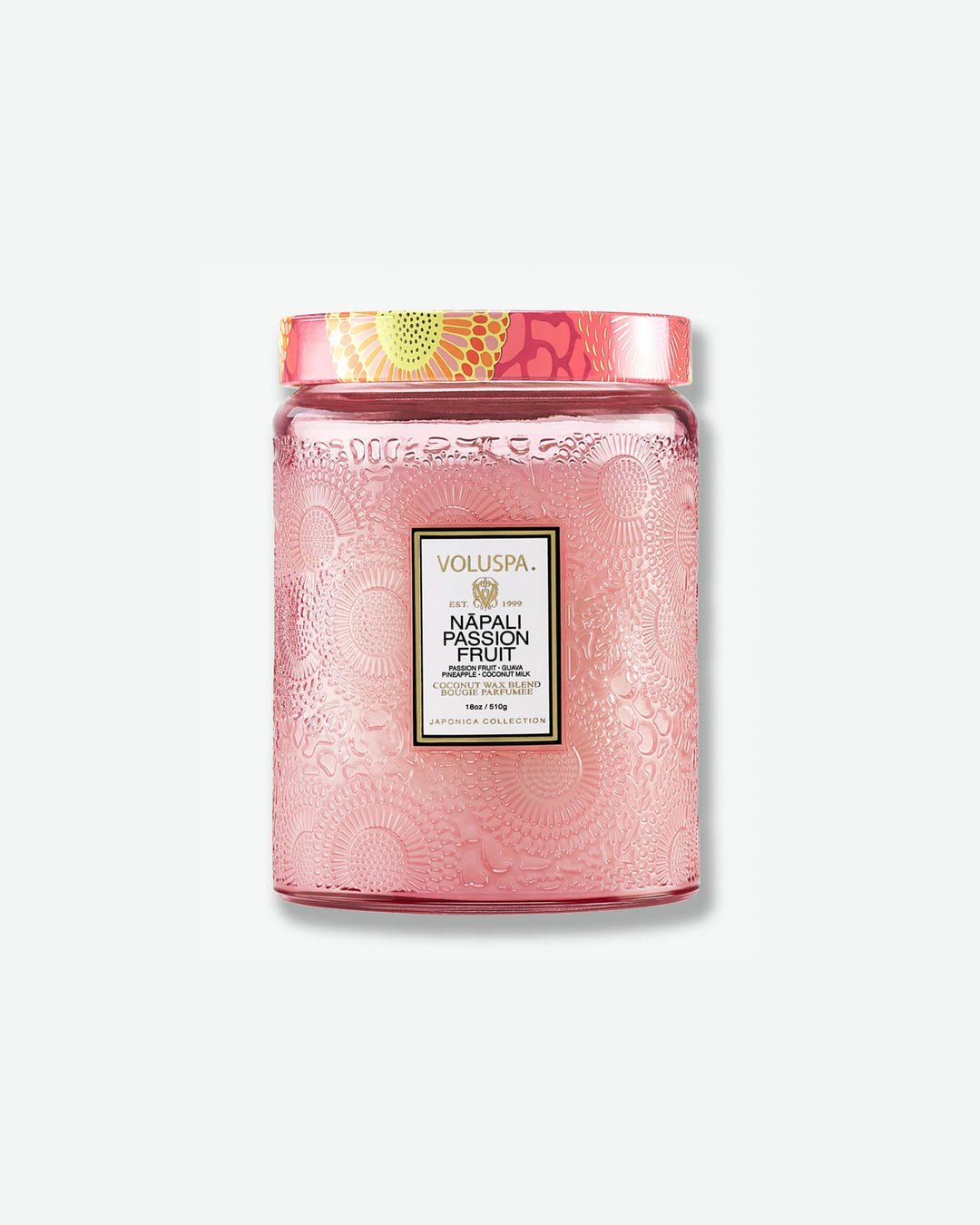 Front view of a Voluspa Nāpali Passion Fruit large jar candle against a plain gray background. The candle is enclosed in a soft pink translucent glass container beautifully embossed with floral motifs, detailed with a central black label and topped with a pink, orange, and yellow patterned tin lid.