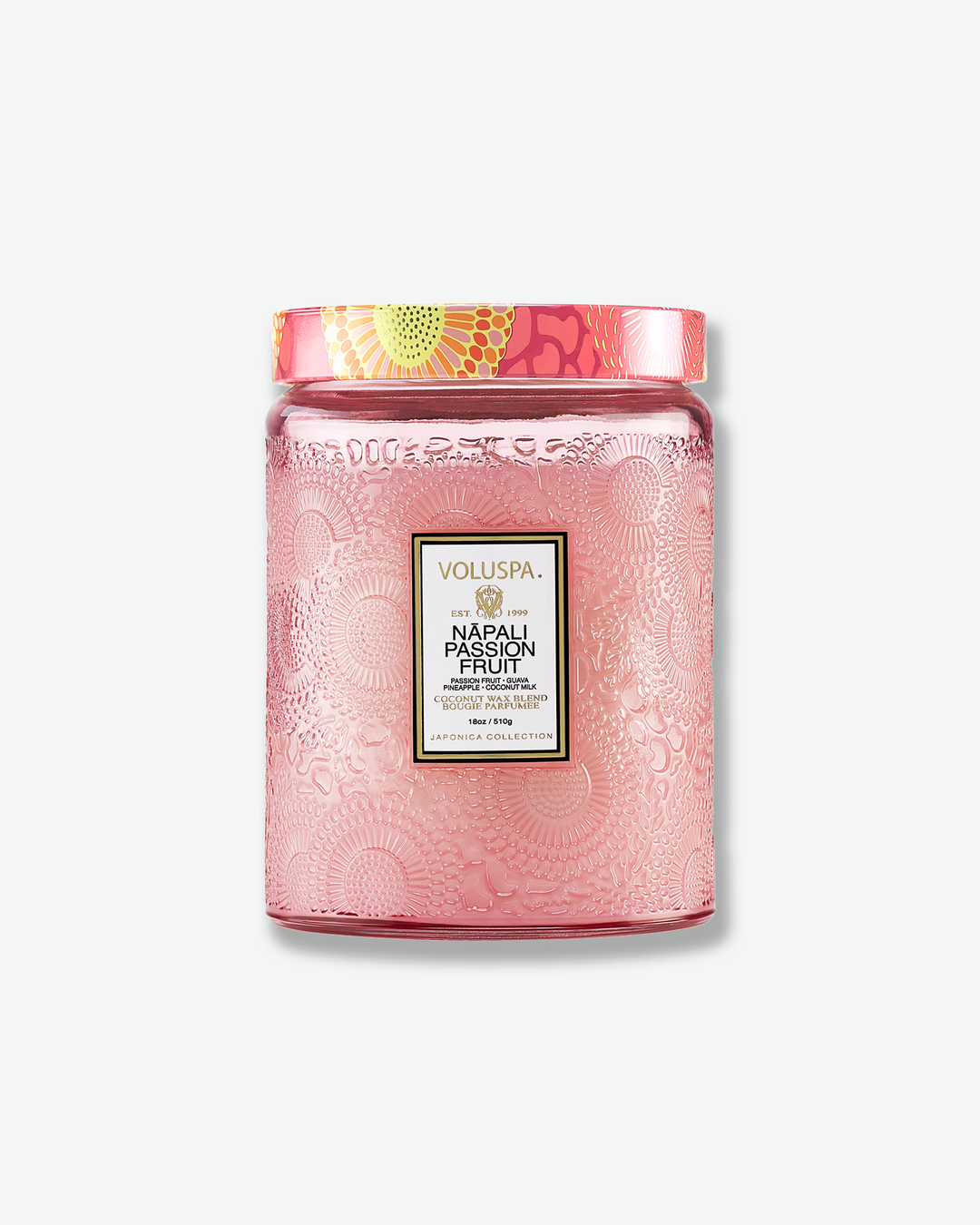 Front view of a Voluspa Nāpali Passion Fruit large jar candle against a plain gray background. The candle is enclosed in a soft pink translucent glass container beautifully embossed with floral motifs, detailed with a central black label and topped with a pink, orange, and yellow patterned tin lid.