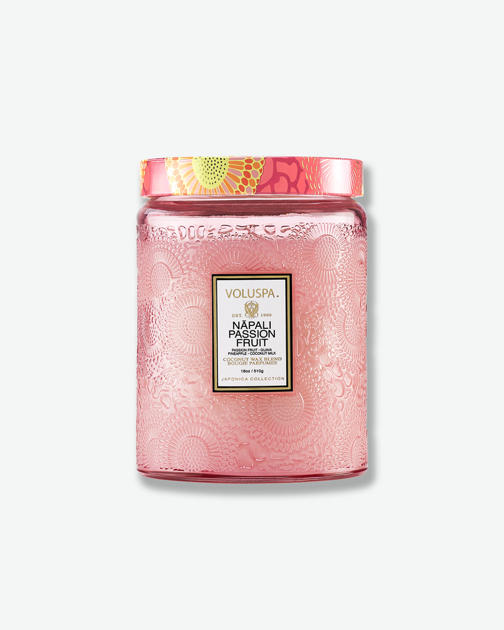 Front view of a Voluspa Nāpali Passion Fruit large jar candle against a plain gray background. The candle is enclosed in a soft pink translucent glass container beautifully embossed with floral motifs, detailed with a central black label and topped with a pink, orange, and yellow patterned tin lid.
