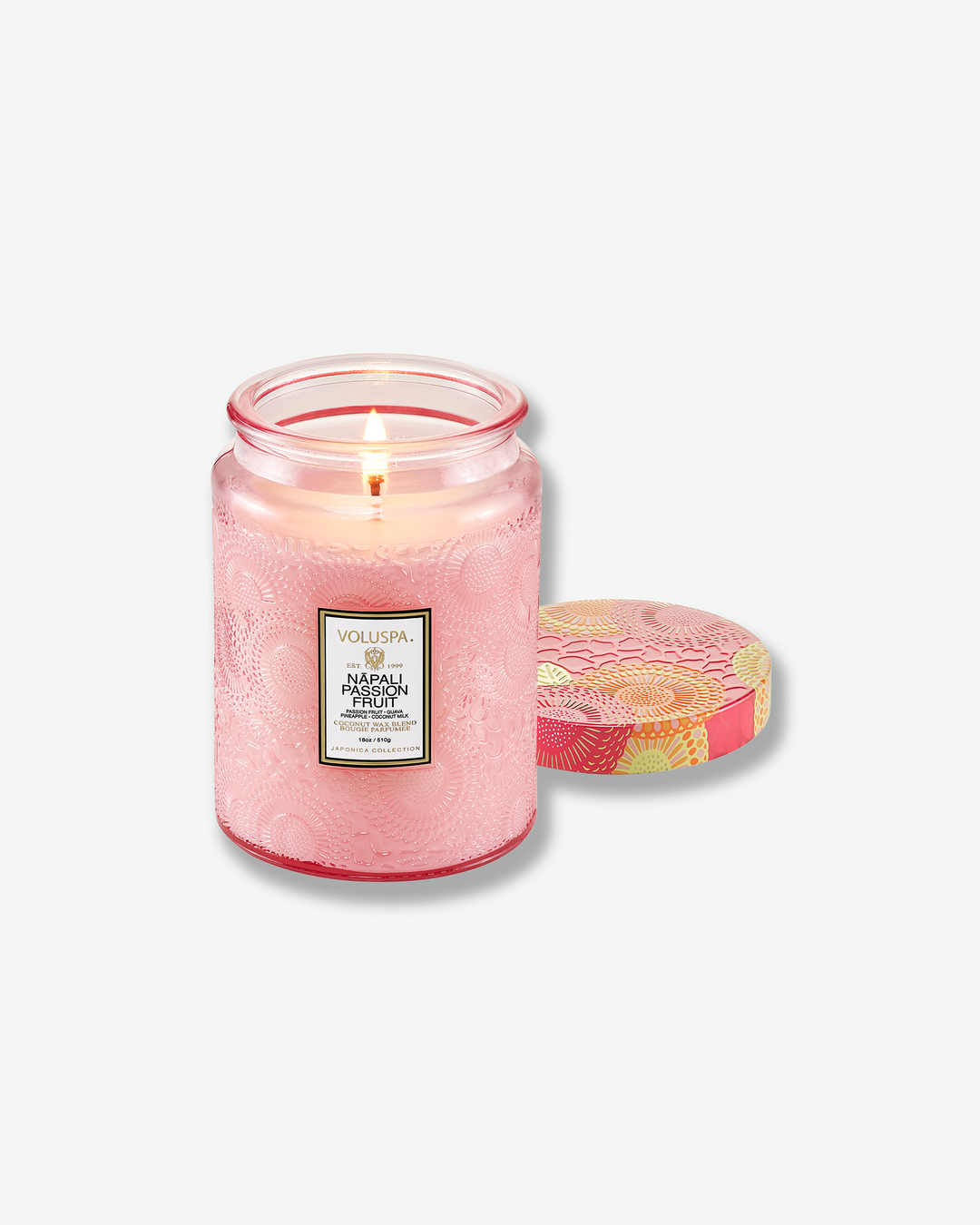 Product shot of a lit Voluspa Nāpali Passion Fruit large jar candle. The single wick is illuminated inside the pink embossed jar, with the decorative multi-colored floral tin lid resting face-up next to the candle.