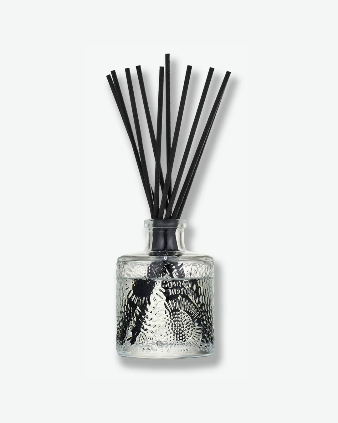 Front view of a Voluspa reed diffuser against a plain gray background. The clear glass bottle is decorated with an intricate embossed floral texture and holds clear fragrance oil, with a collection of long black reeds extending upward from the narrow neck.