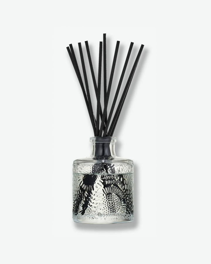 Front view of a Voluspa reed diffuser against a plain gray background. The clear glass bottle is decorated with an intricate embossed floral texture and holds clear fragrance oil, with a collection of long black reeds extending upward from the narrow neck.
