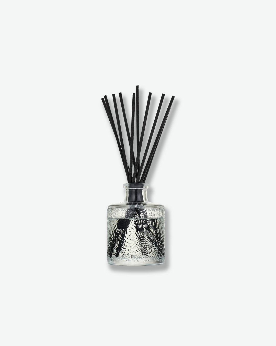Front view of a Voluspa reed diffuser against a plain gray background. The clear glass bottle is decorated with an intricate embossed floral texture and holds clear fragrance oil, with a collection of long black reeds extending upward from the narrow neck.