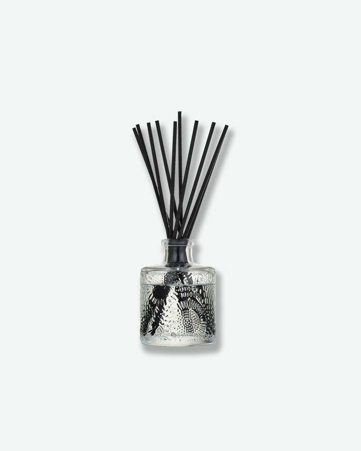 Front view of a Voluspa reed diffuser against a plain gray background. The clear glass bottle is decorated with an intricate embossed floral texture and holds clear fragrance oil, with a collection of long black reeds extending upward from the narrow neck.