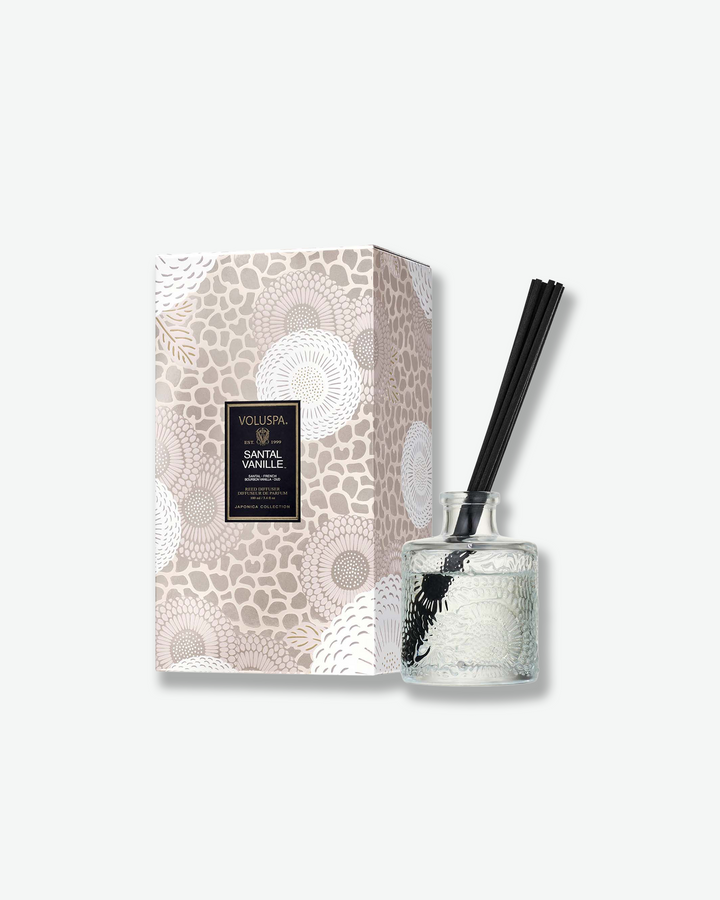 Product shot featuring a Voluspa Santal Vanille reed diffuser standing next to its decorative packaging box against a plain gray background. The clear, embossed glass bottle with a bundle of black reeds sits beside a vertical rectangular gift box adorned with a soft beige and white botanical floral pattern.