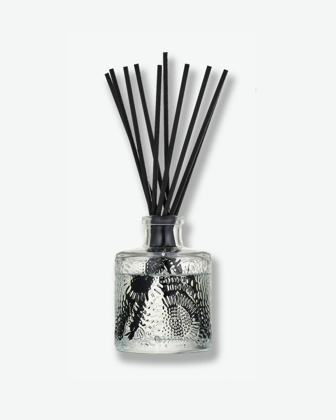 Front view of a Voluspa reed diffuser bottle against a plain gray background. The small, cylindrical clear glass container features an intricate, embossed floral texture and holds a bundle of long black diffuser reeds extending outward from its narrow neck.