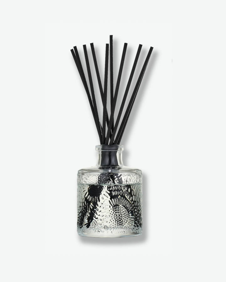 Front view of a Voluspa reed diffuser bottle against a plain gray background. The small, cylindrical clear glass container features an intricate, embossed floral texture and holds a bundle of long black diffuser reeds extending outward from its narrow neck.