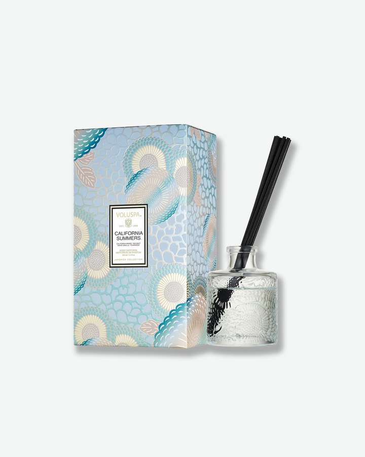 Product shot featuring a Voluspa Mediterranean Lemon reed diffuser standing next to its decorative packaging box. The clear, embossed glass diffuser bottle with black reeds sits to the right of a vertical rectangular gift box adorned with a cobalt blue, gold, and white botanical pattern.