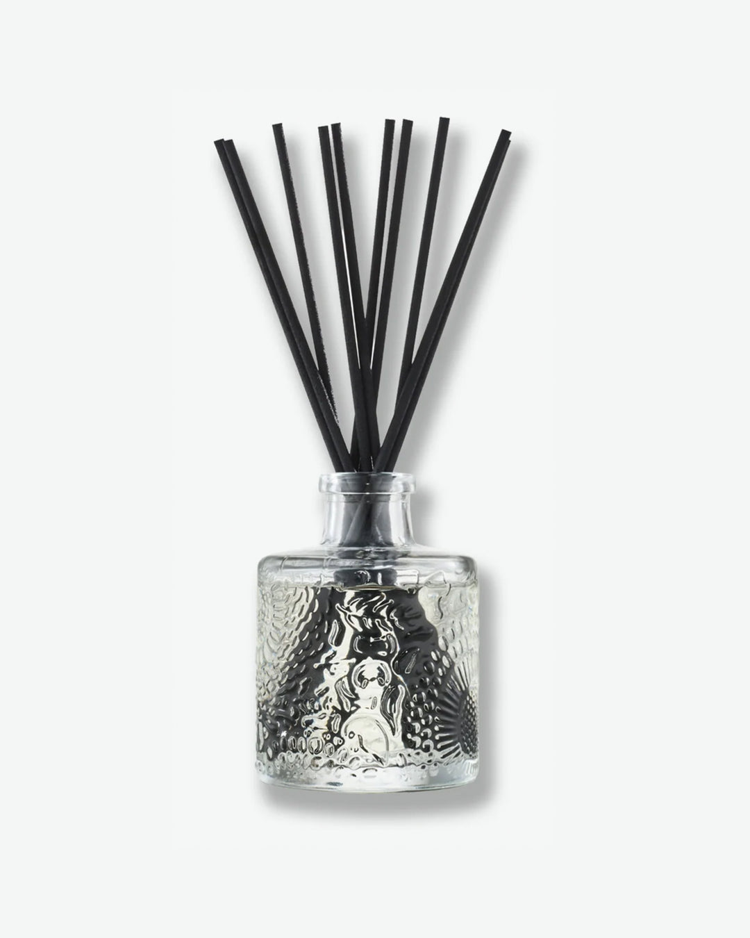 Front view of a Voluspa reed diffuser against a plain gray background. The clear glass bottle is detailed with an intricate embossed floral texture and holds a collection of long black reeds extending upward from the narrow neck.