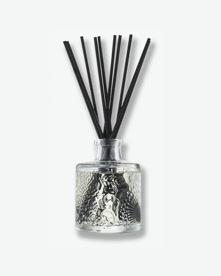 Front view of a Voluspa reed diffuser against a plain gray background. The clear glass bottle is detailed with an intricate embossed floral texture and holds a collection of long black reeds extending upward from the narrow neck.