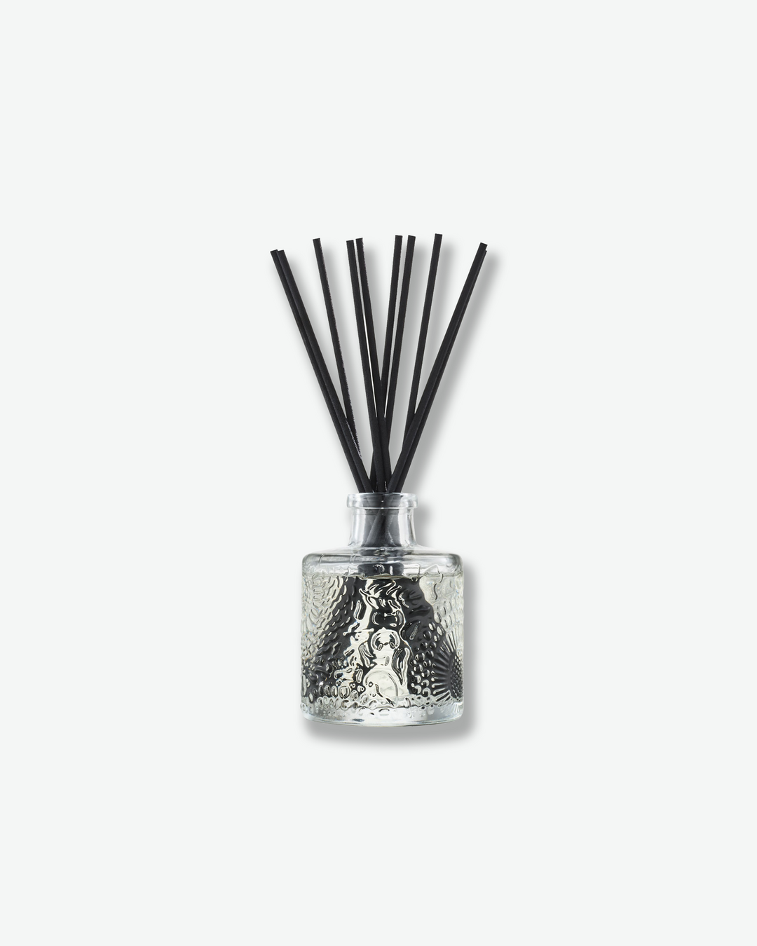 Front view of a Voluspa reed diffuser against a plain gray background. The clear glass bottle is detailed with an intricate embossed floral texture and holds a collection of long black reeds extending upward from the narrow neck.