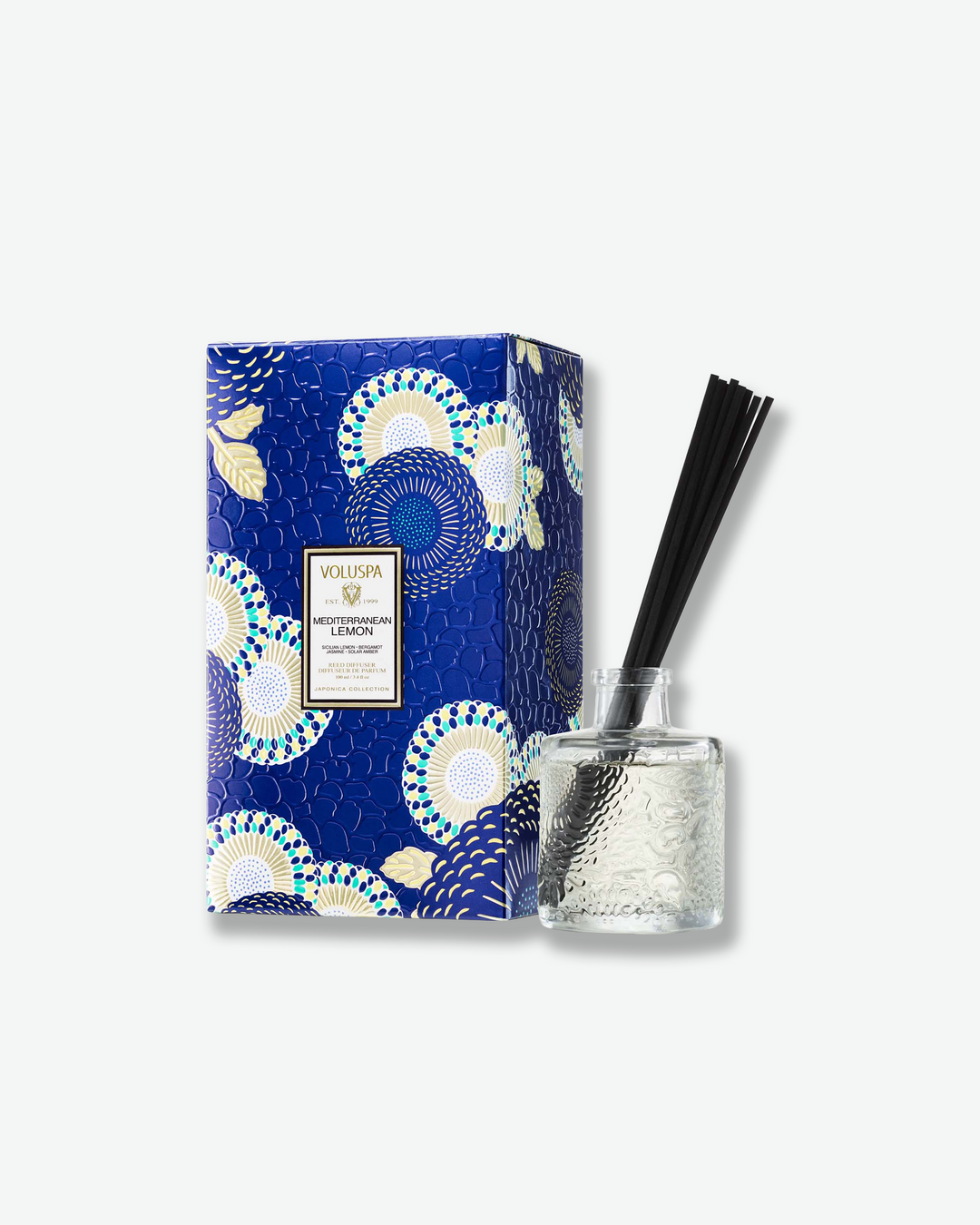 Product shot featuring a Voluspa California Summers reed diffuser standing next to its decorative packaging box against a plain gray background. The clear, embossed glass bottle with a bundle of black reeds sits beside a vertical rectangular gift box adorned with a light icy-blue and silver botanical pattern.