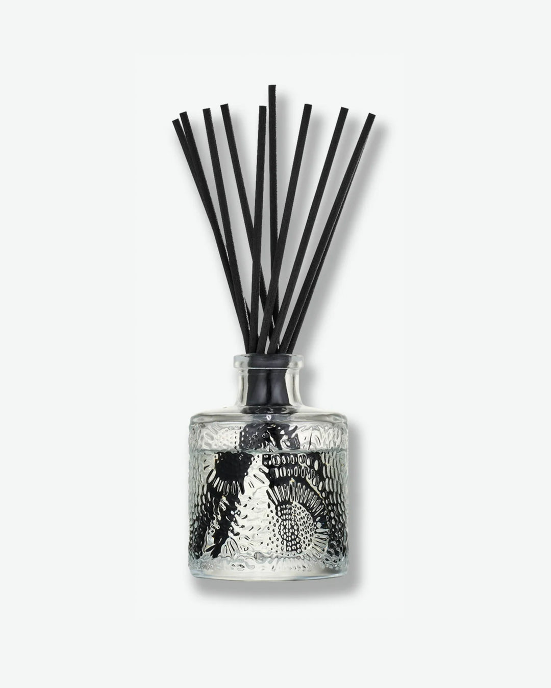 Front view of a Voluspa reed diffuser against a plain gray background. The clear glass vessel is detailed with a swirling, embossed floral pattern and filled with fragrance fluid, holding a collection of long black reeds fanning out from the top.