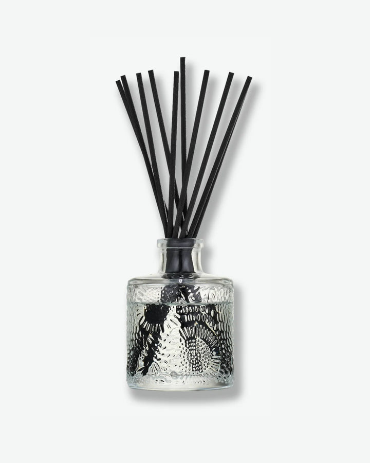 Front view of a Voluspa reed diffuser against a plain gray background. The clear glass vessel is detailed with a swirling, embossed floral pattern and filled with fragrance fluid, holding a collection of long black reeds fanning out from the top.