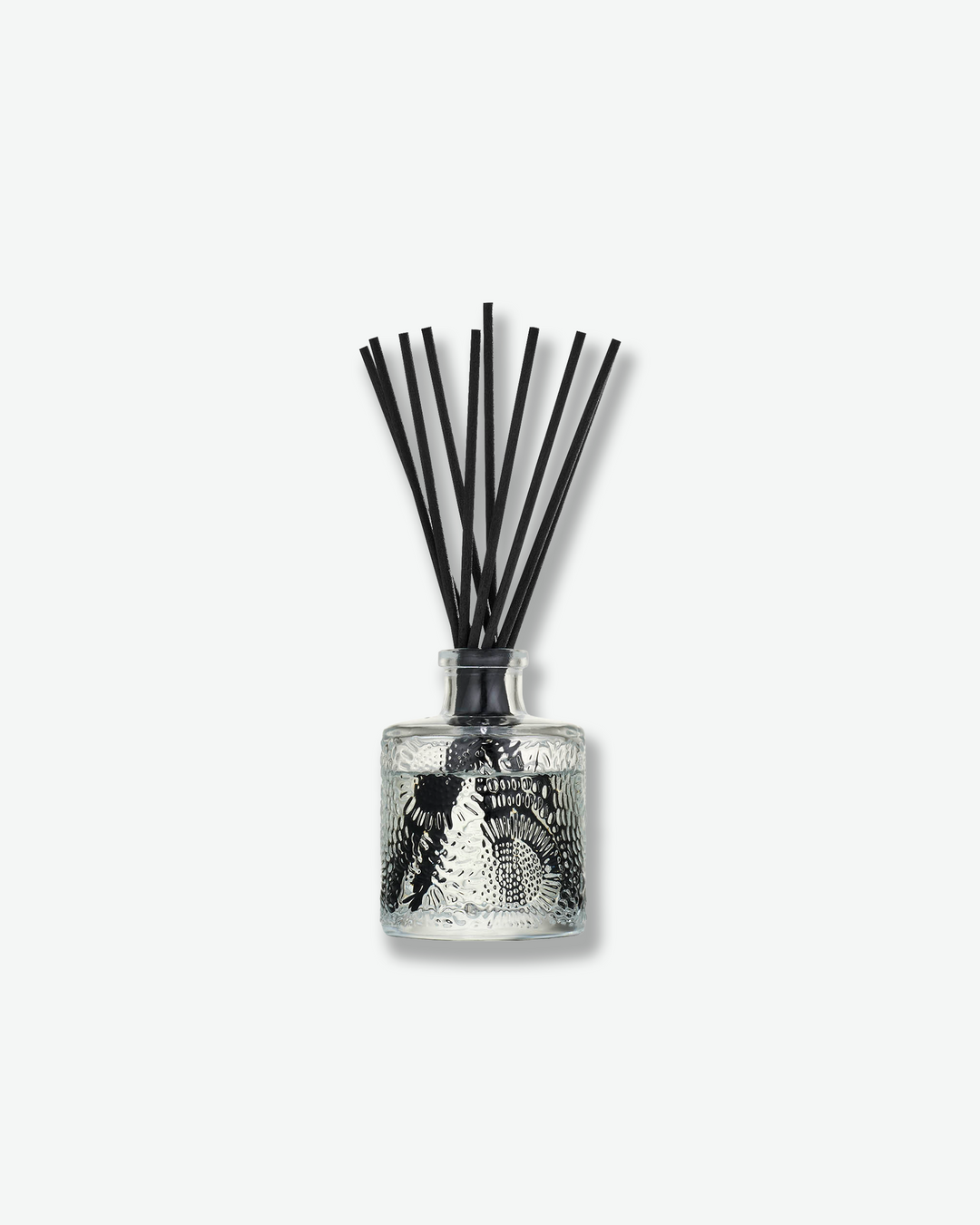 Front view of a Voluspa reed diffuser against a plain gray background. The clear glass vessel is detailed with a swirling, embossed floral pattern and filled with fragrance fluid, holding a collection of long black reeds fanning out from the top.