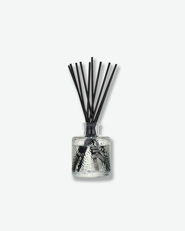 Front view of a Voluspa reed diffuser against a plain gray background. The clear glass vessel is detailed with a swirling, embossed floral pattern and filled with fragrance fluid, holding a collection of long black reeds fanning out from the top.