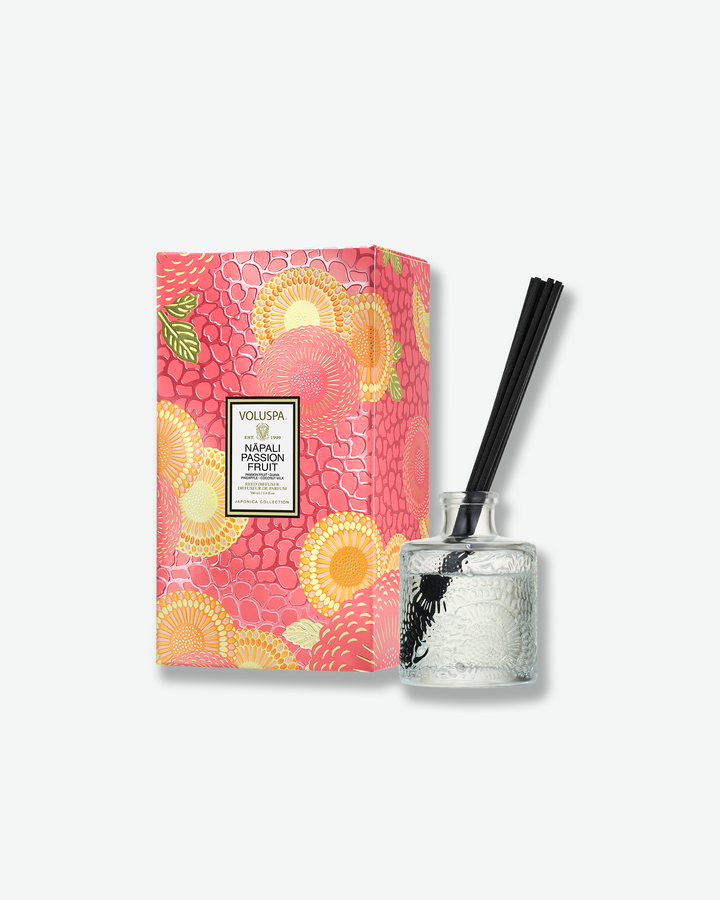 Product shot showcasing a Voluspa Nāpali Passion Fruit reed diffuser paired with its matching packaging box. The clear, embossed glass bottle with a few black reeds stands beside a vertical rectangular gift box styled with a bright pink, orange, and yellow botanical design.