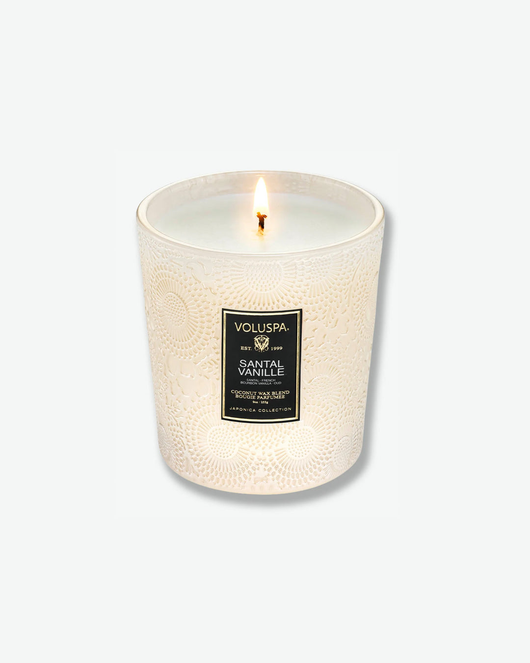 High-angle product shot of a burning Voluspa Santal Vanille classic textured glass candle against a plain gray background. The single-wick flame glows from the center of the pool of melted white wax inside the elegant, cream-colored embossed glass holder.