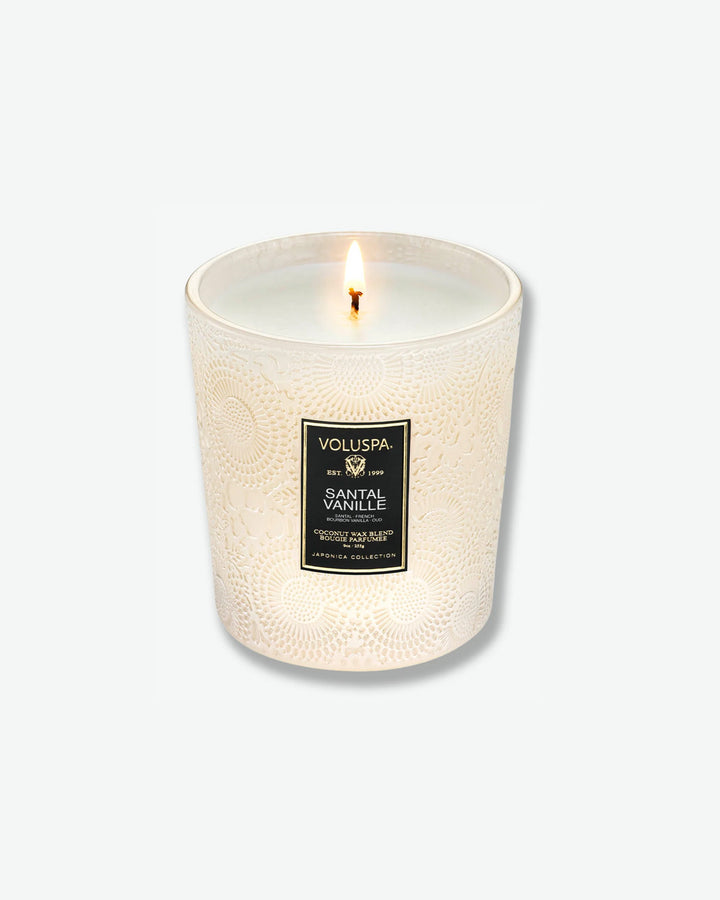 High-angle product shot of a burning Voluspa Santal Vanille classic textured glass candle against a plain gray background. The single-wick flame glows from the center of the pool of melted white wax inside the elegant, cream-colored embossed glass holder.