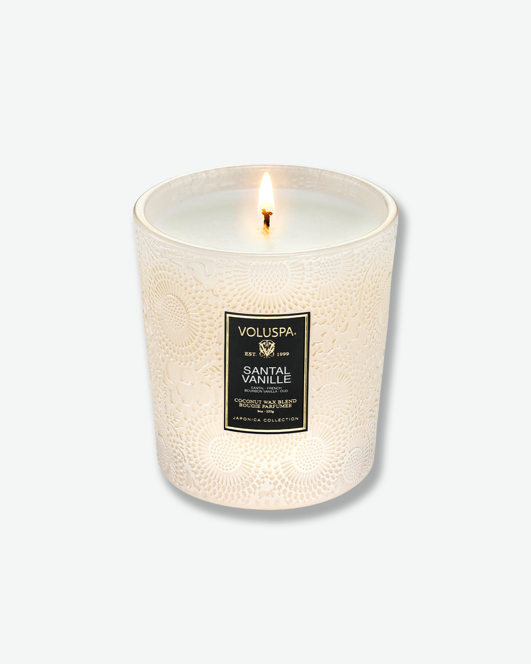 High-angle product shot of a burning Voluspa Santal Vanille classic textured glass candle against a plain gray background. The single-wick flame glows from the center of the pool of melted white wax inside the elegant, cream-colored embossed glass holder.