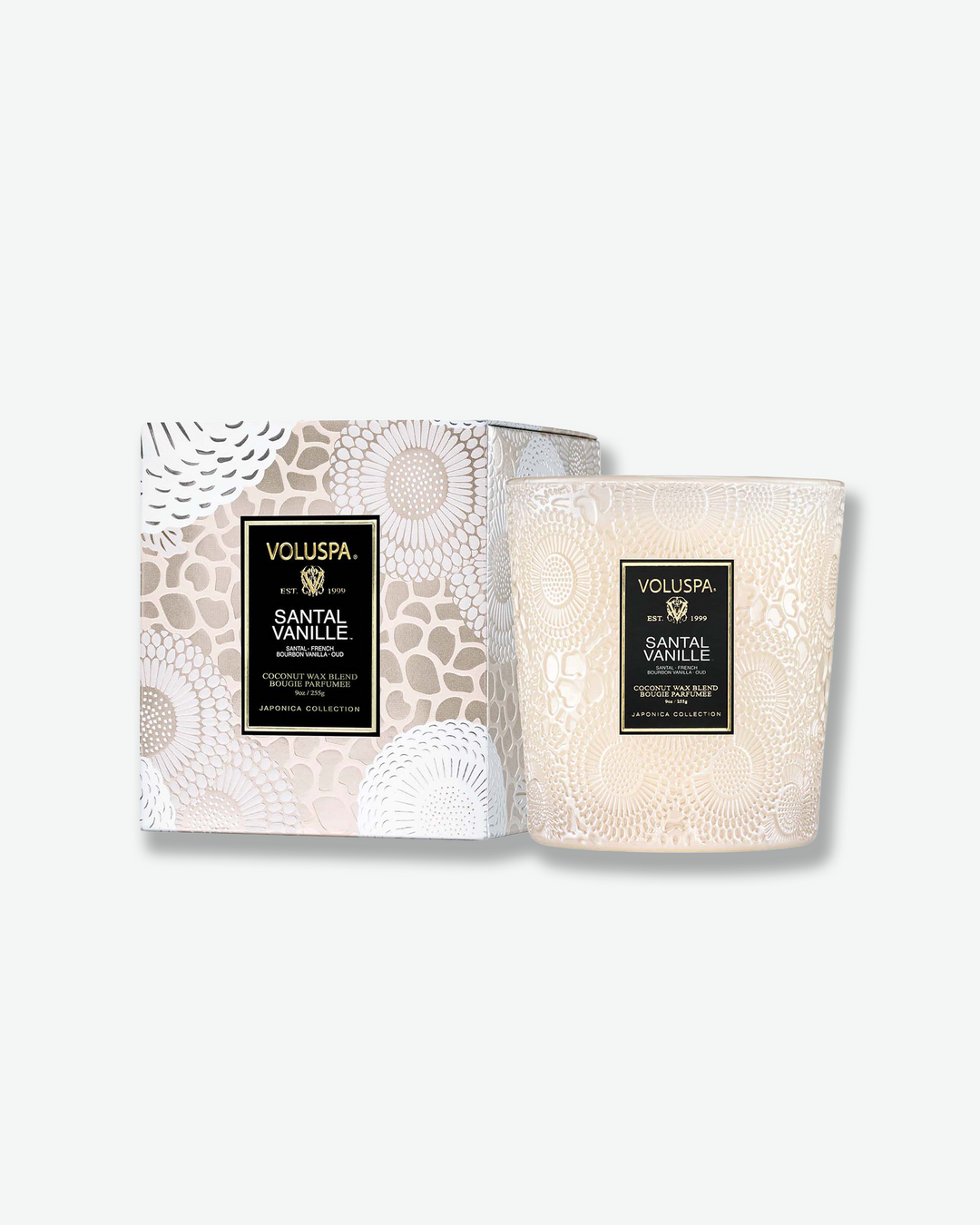Product shot featuring a Voluspa Santal Vanille classic textured glass candle next to its square outer box. The cream-tinted glass candle vessel features a tapered shape and sits beside a box patterned with a matching muted beige and white Japonica floral design.