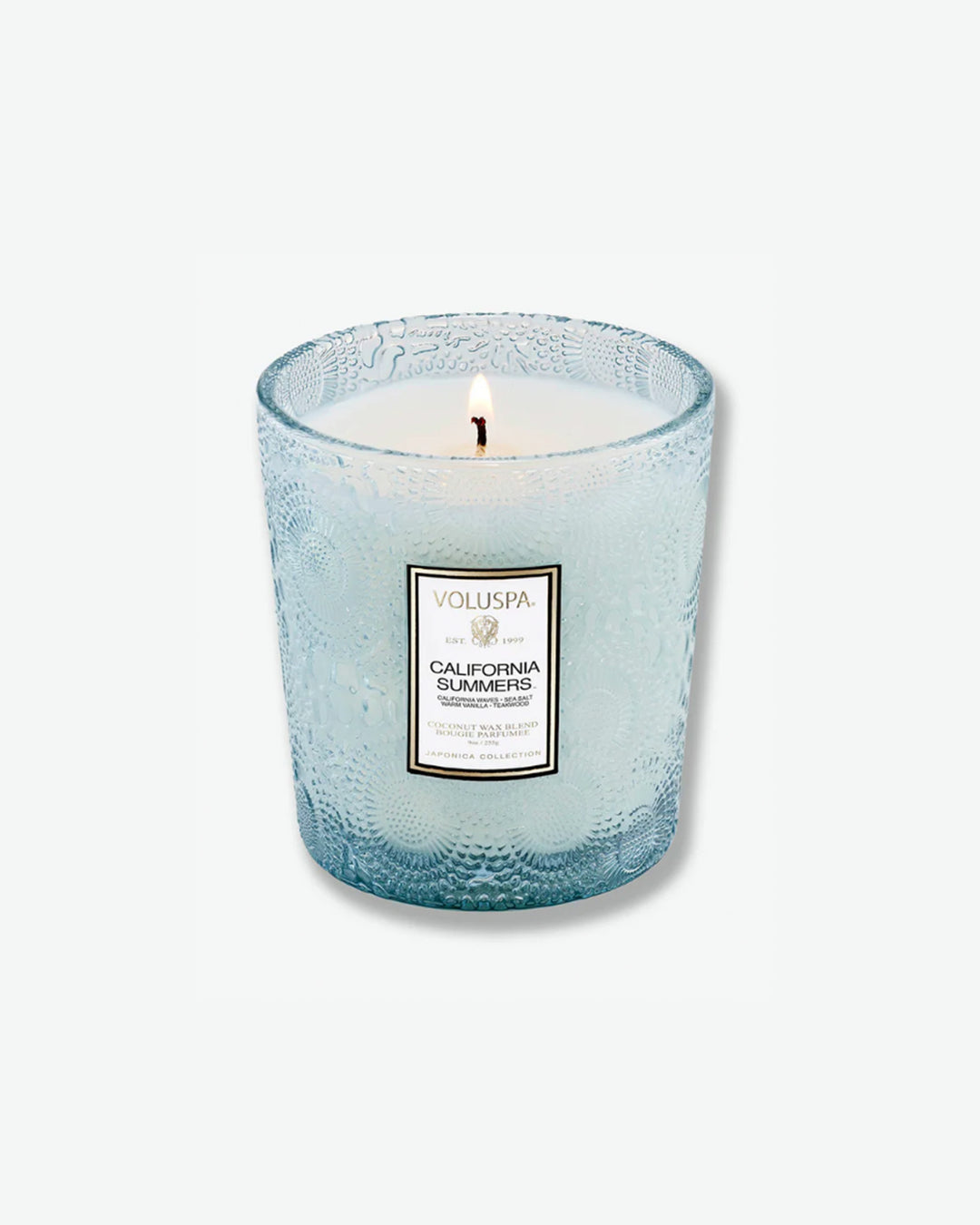 Product shot of a Voluspa California Summers classic textured glass candle standing next to its matching square outer packaging. The light blue glass candle jar is detailed with an intricate embossed floral motif and sits beside a box featuring a light blue, cream, and silver botanical pattern.