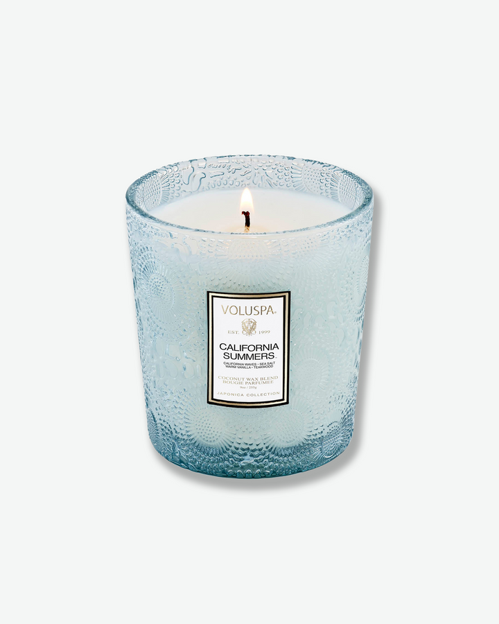 Product shot of a Voluspa California Summers classic textured glass candle standing next to its matching square outer packaging. The light blue glass candle jar is detailed with an intricate embossed floral motif and sits beside a box featuring a light blue, cream, and silver botanical pattern.