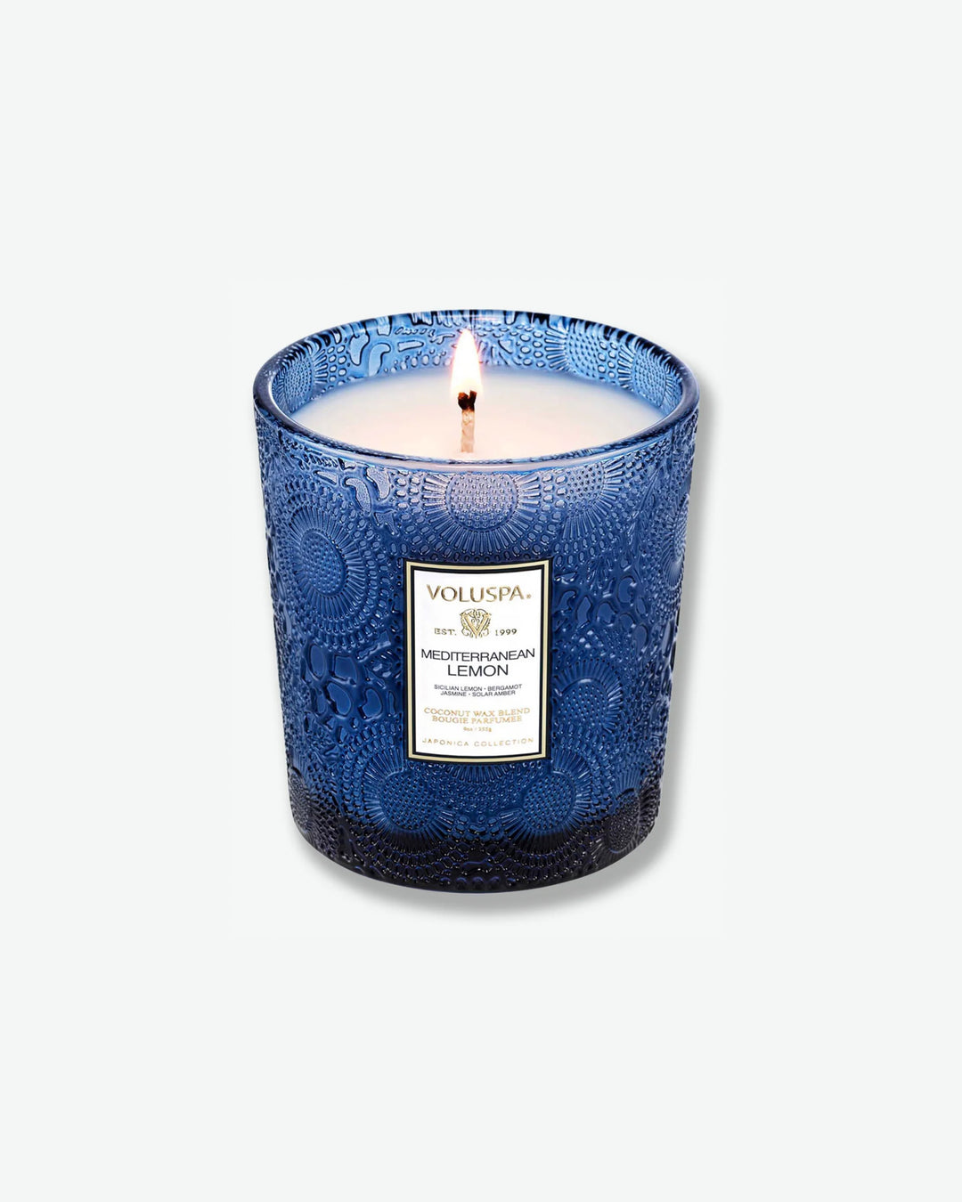 High-angle product shot of a burning Voluspa Mediterranean Lemon classic textured glass candle against a plain gray background. The single-wick flame burns in the center of the deep cobalt blue, embossed glass holder, illuminating the pool of melted white wax.