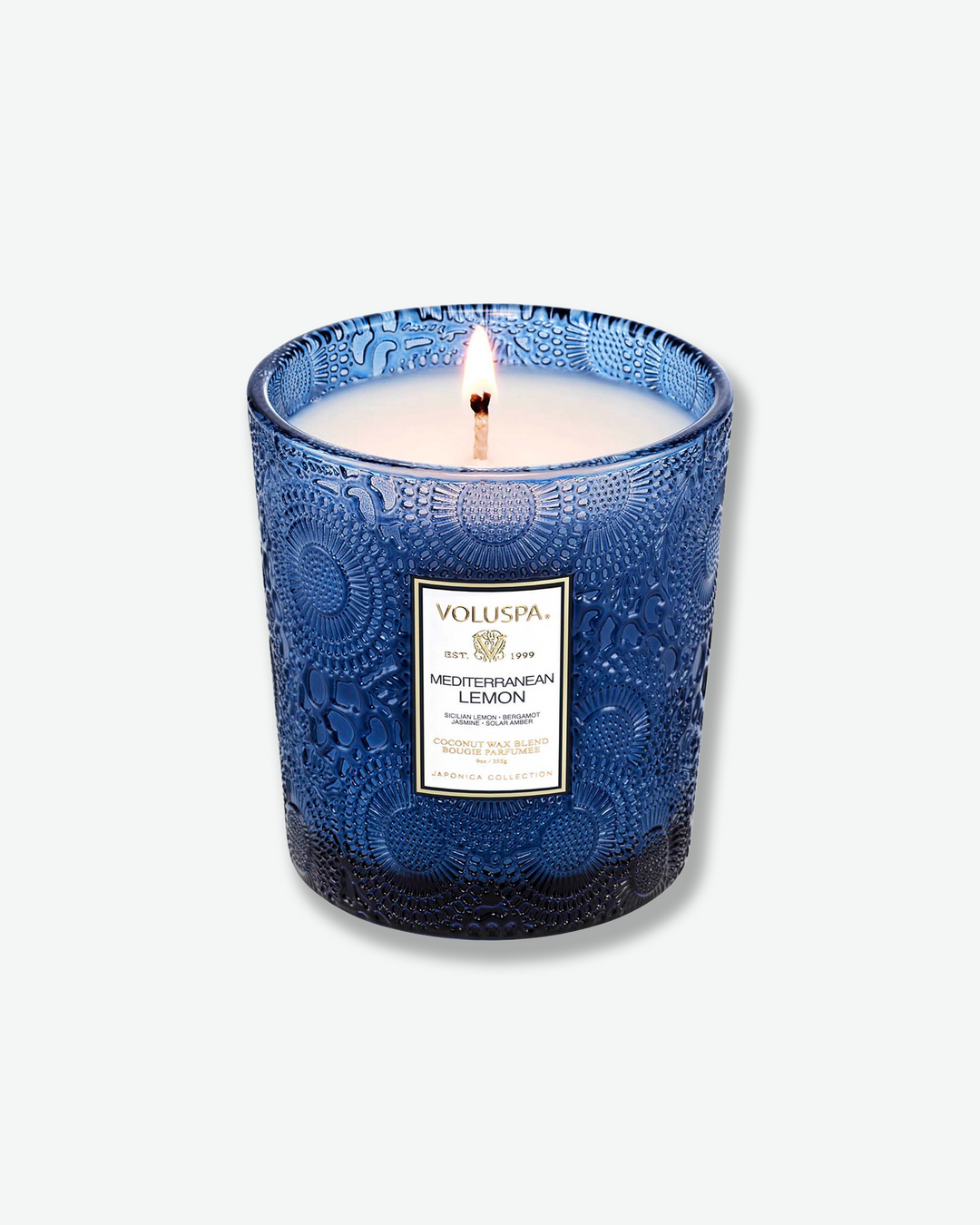 High-angle product shot of a burning Voluspa Mediterranean Lemon classic textured glass candle against a plain gray background. The single-wick flame burns in the center of the deep cobalt blue, embossed glass holder, illuminating the pool of melted white wax.