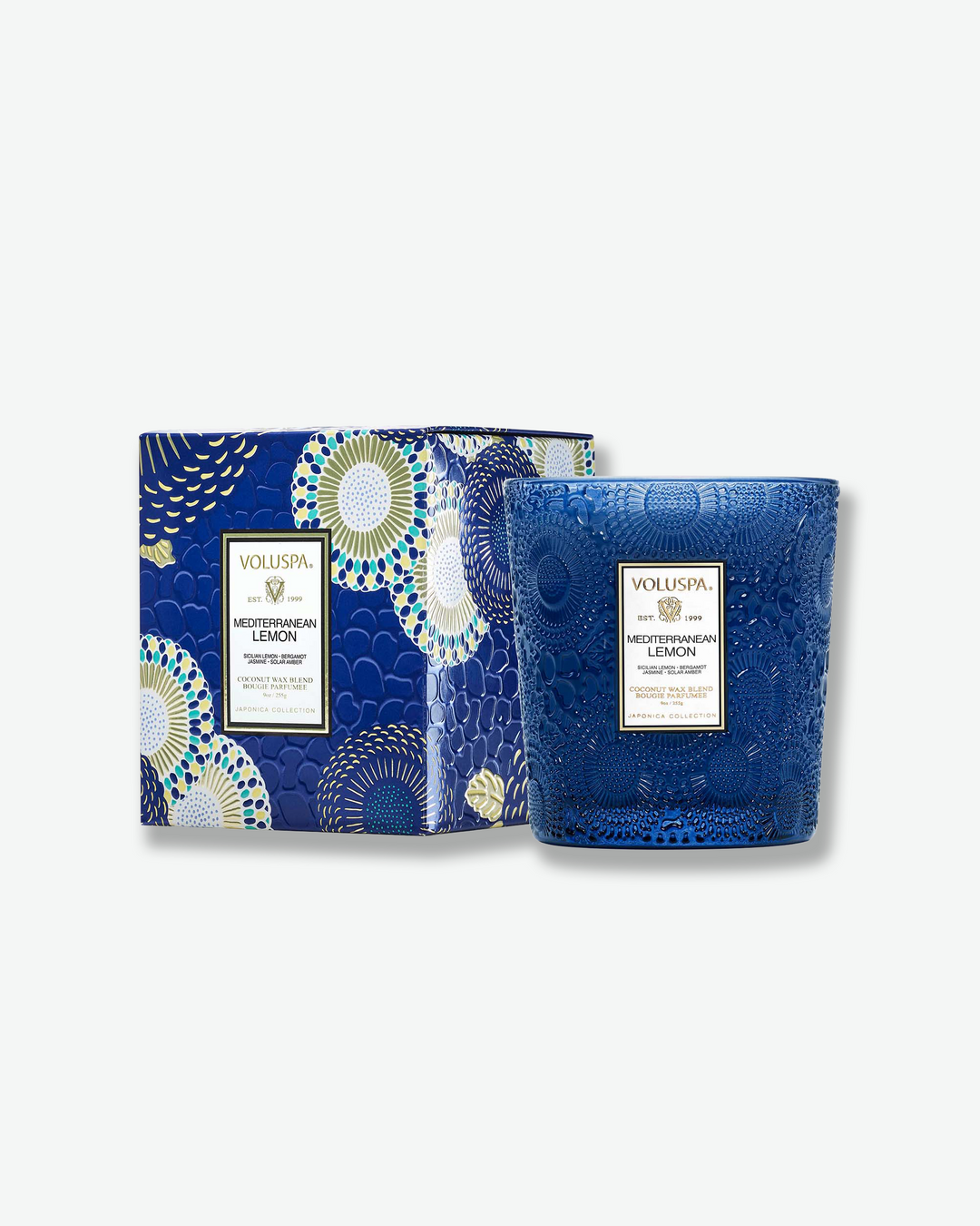 Product shot showcasing a Voluspa Mediterranean Lemon classic textured glass candle next to its square outer box. The deep cobalt blue glass candle vessel features a tapered shape and sits beside a box patterned with a matching blue, gold, and white Japonica floral design.